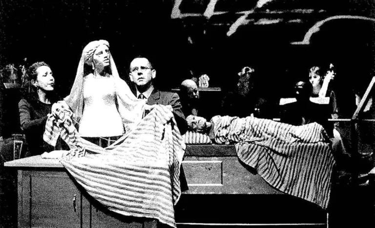 A l’avant-plan Pénélope file la laine dans la couverture-costume d’Ulysse Guillaumette Laurens, chanteuse, Adrian Kohler, acteur-manipulateur. À l’arrière-plan Ulysse, alité est soutenu par Scott Weir, chanteur, Basil Jones, acteur-manipulateur et les musiciens. Photo Herman Sorgeloos.